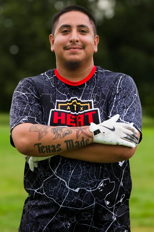 An ATX Heat player poses with arms crossed on a grassy field during Gay Bowl 2025 practice, wearing a black jersey with a white map pattern, red collar, and an “ATX HEAT” logo. The athlete has white football gloves and stands in an outdoor park setting with blurred trees in the background.