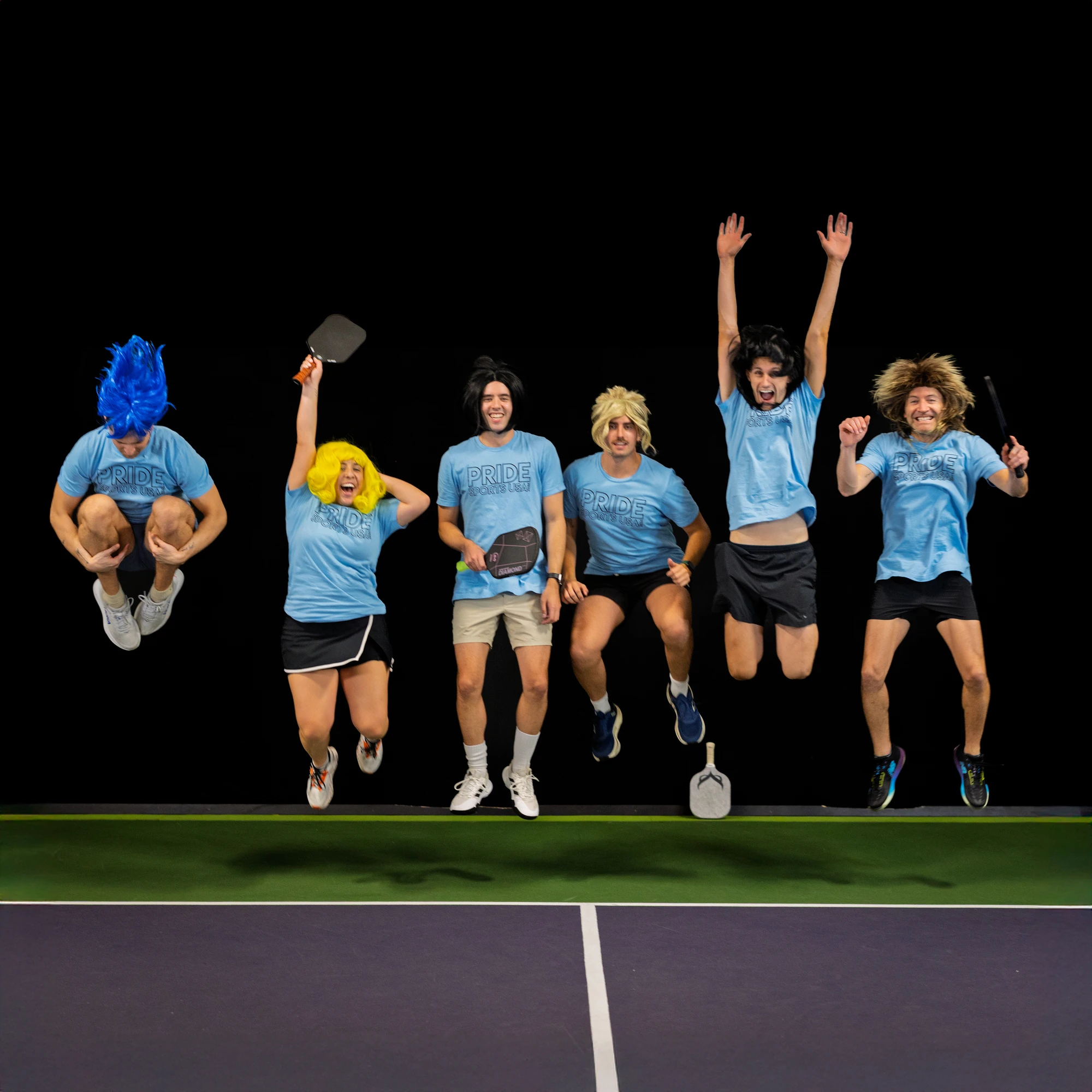 A group of pickleball players wearing colorful wigs and matching blue "Pride" shirts are on a pickleball court, energetically jumping in the air and holding their paddles.