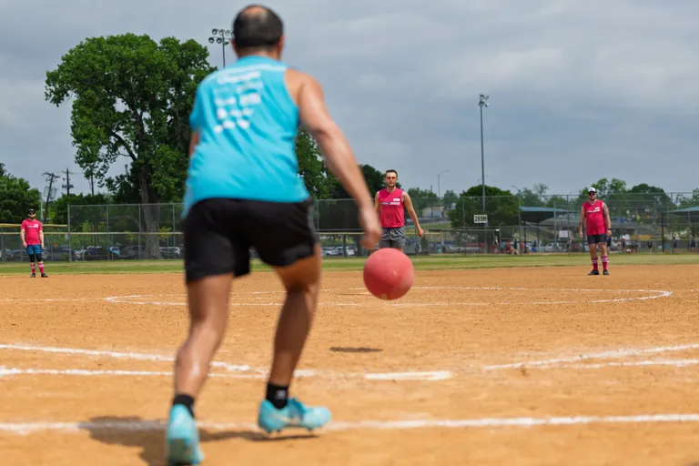 A player in a blue tank top and black shorts is running on a kickball field, preparing to kick a red kickball, while several other players in pink shirts stand ready in the outfield. The scene takes place outdoors on a dirt field with a fence, trees, and sports lights visible in the background.