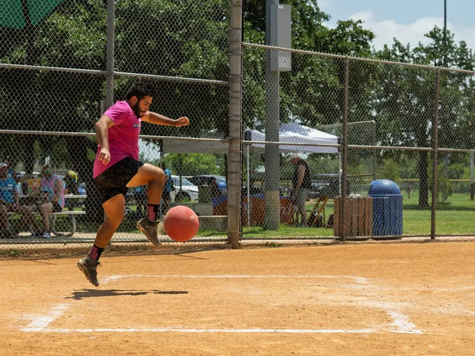 A player wearing a bright pink shirt and black shorts is in mid-air preparing to kick a red kickball on a sandy kickball field, with spectators and a chain-link fence in the background.