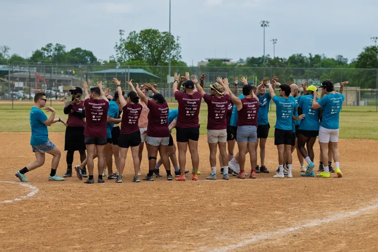 Players wearing maroon and teal team shirts are gathering in a circle on a kickball field, raising their arms and celebrating together after a game. The field is surrounded by grass, fencing, and distant sports facilities.
