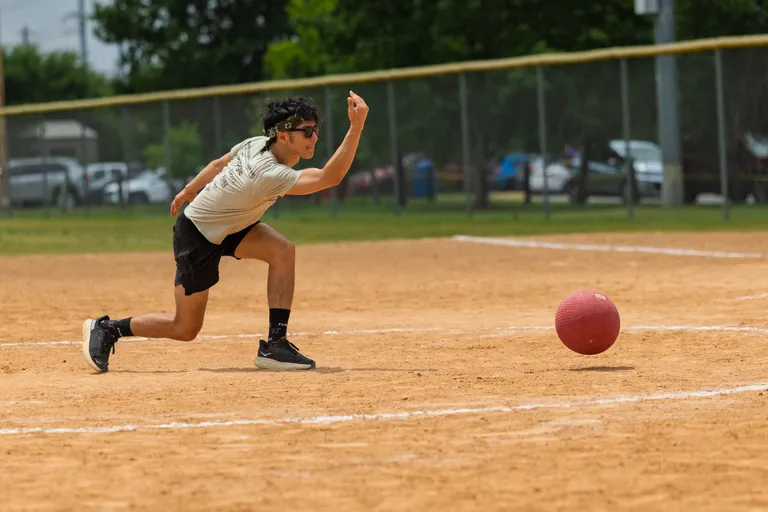 A kickball player wearing a light shirt, dark shorts, and a headband is in mid-throw on a dirt kickball field, with a red kickball rolling near the pitcher's mound.