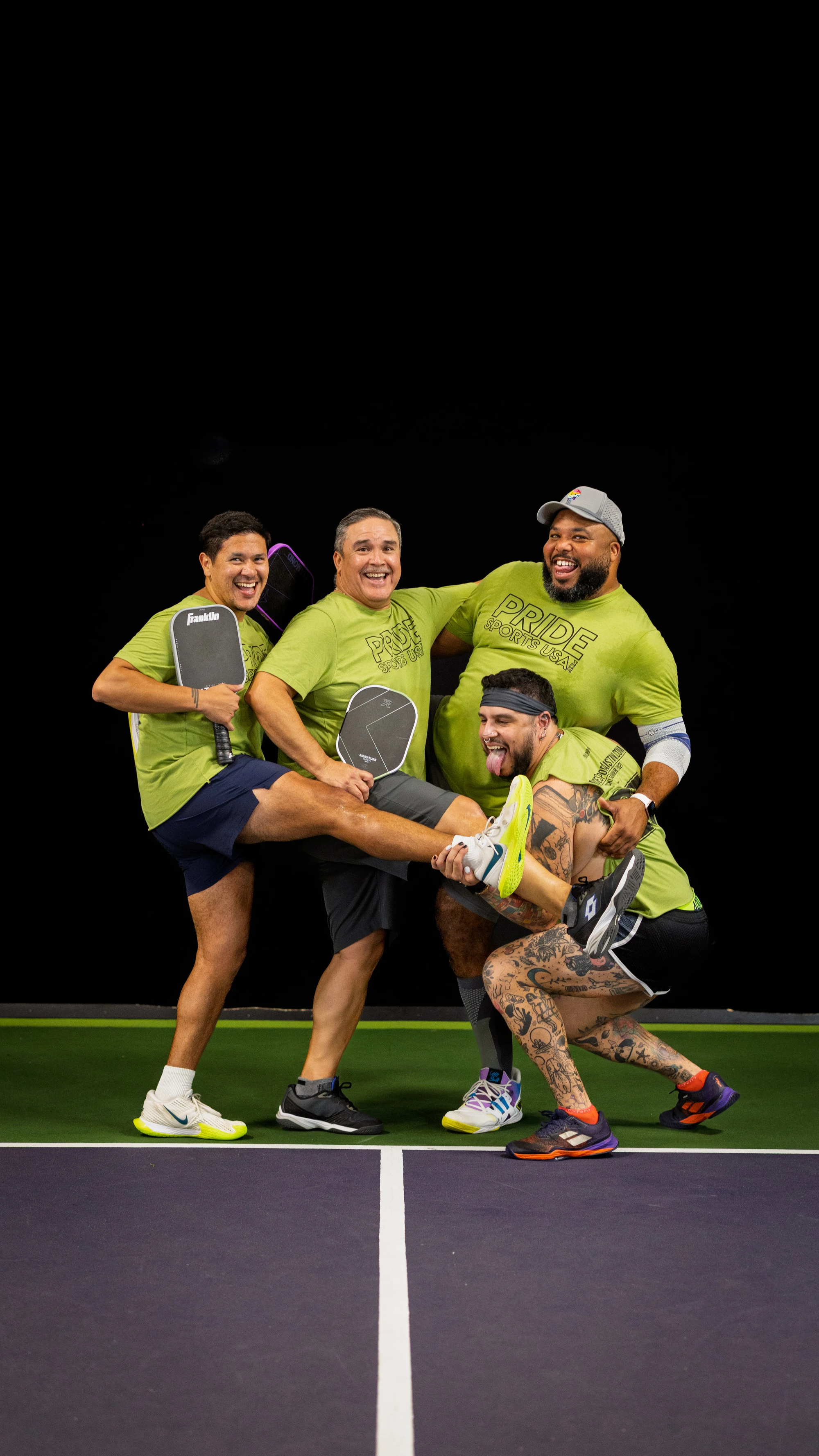 Four pickleball players wearing matching lime green "Pride" t-shirts pose playfully on an indoor court, holding paddles and laughing together, with one player crouched at the front and sticking out their tongue while the group supports each other in a lively, celebratory stance.