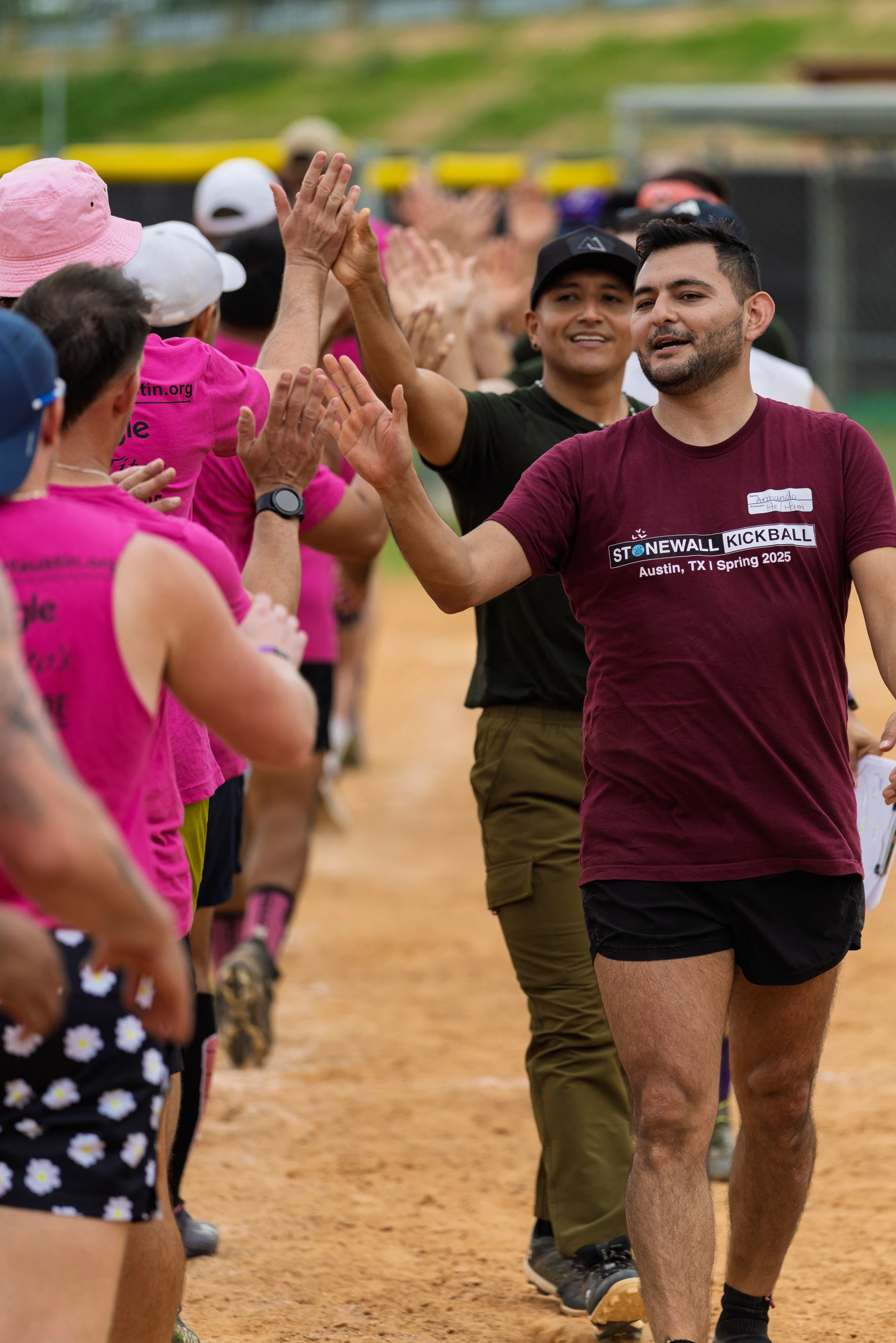 Players wearing team shirts, including one in a maroon "Stonewall Kickball Austin, TX | Spring 2025" shirt, are giving high-fives in two lines on a kickball field, celebrating after a game. The setting is an outdoor dirt field, and the players are dressed in athletic clothing, with some wearing hats and shorts.