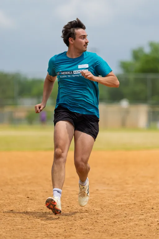 A kickball player wearing a teal "Stonewall Kickball" shirt, black shorts, and white athletic shoes is running across a dirt field, looking to the side during a game. The outdoor setting shows a blurred background of trees and fencing.
