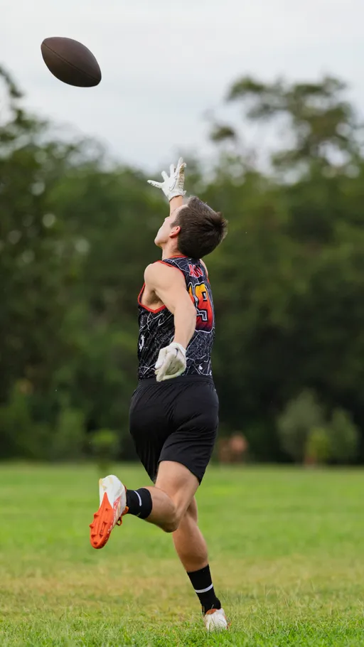 ATX Heat (Orange) player sprints across a grassy field during flag football practice, eyes up as a football drops overhead and white receiver-gloved hands reach to catch it. The athlete wears a black patterned sleeveless jersey with orange trim and numbers, black shorts, and cleats with orange soles, with trees in the background.