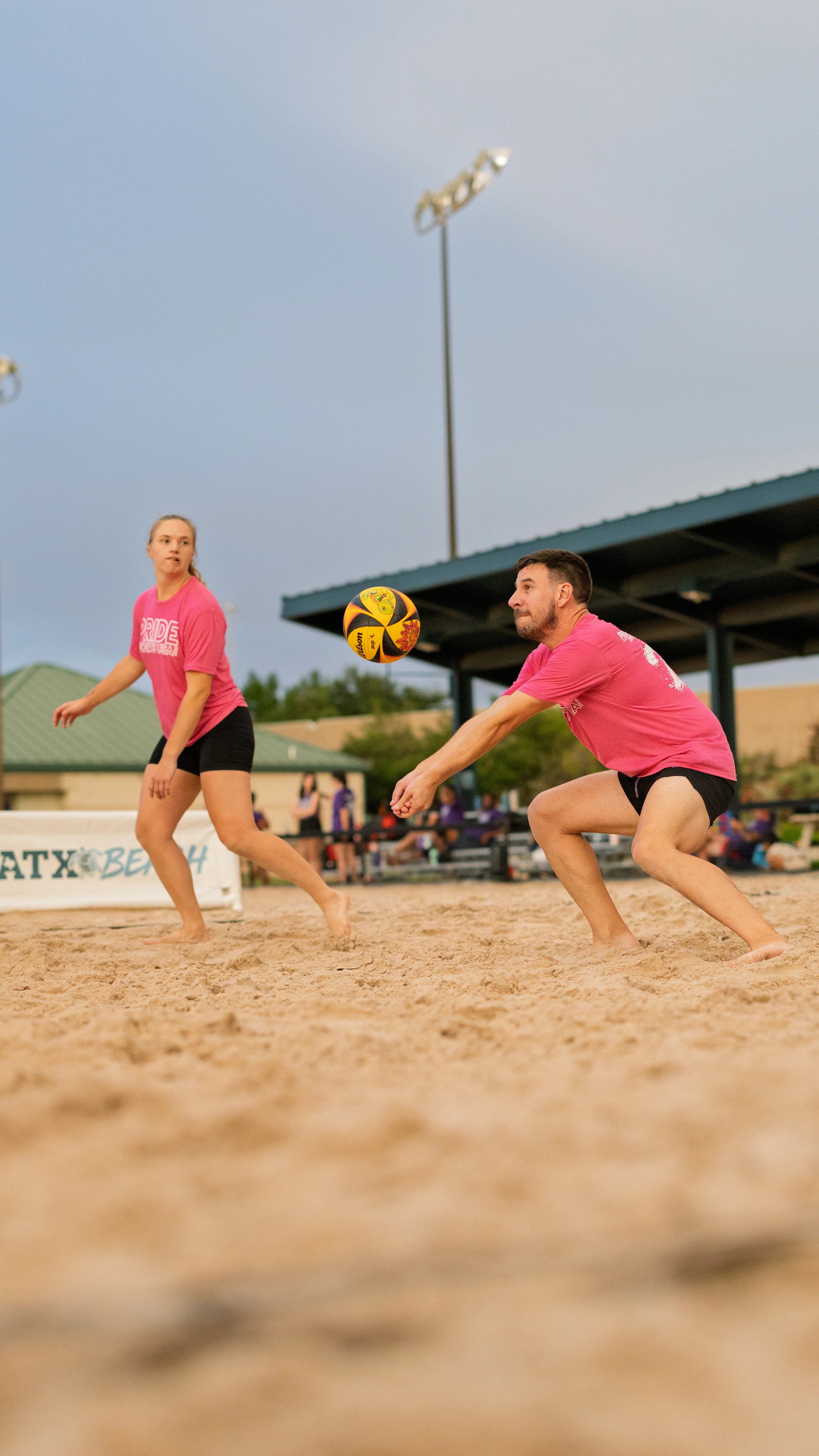 Two volleyball players in matching pink shirts and black shorts are on a sandy court, with one player crouched and arms outstretched to bump the ball while the other watches attentively. Outdoor lights and a covered seating area are visible in the background.