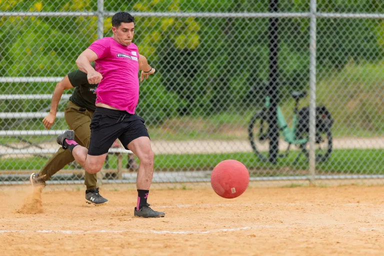 A player in a bright pink shirt and black shorts is kicking a red kickball on a dirt field, while another player in an olive green shirt runs behind them; a chain-link fence and a bicycle are visible in the background.