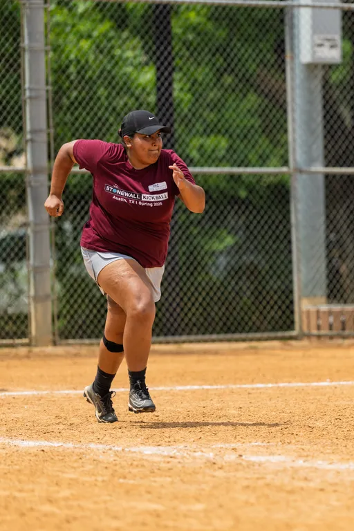 A player in a maroon Stonewall Kickball t-shirt, athletic shorts, cap, and sneakers is running on a kickball field, with a chain-link fence and trees in the background.