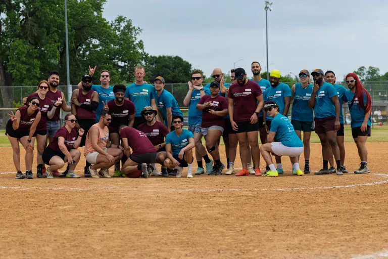 A group of kickball players wearing matching maroon and blue team shirts pose together on a dirt kickball field, some kneeling and some standing, with trees and fencing visible in the background.