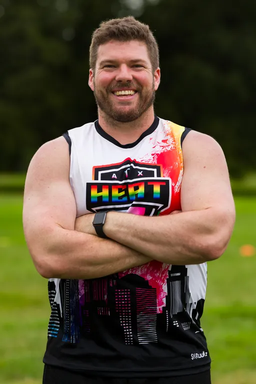 An ATX Heat player stands on a grassy football practice field, smiling with arms crossed, wearing a sleeveless team jersey with rainbow “HEAT” text, cityscape graphics, and an orange shoulder panel, plus a fitness watch; blurred cones mark the field in the background.