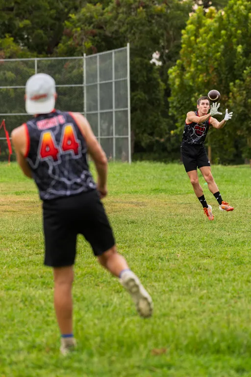 On a grassy park field, an ATX Heat (Orange) quarterback in a black-and-orange web-pattern jersey throws to a receiver sprinting forward, hands up in white gloves as the football arcs toward them. Players wear matching sleeveless jerseys (number 44 visible), shorts, and cleats, with a chain-link backstop and trees in the background during flag football practice for Gay Bowl 2025.