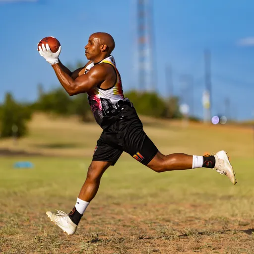 An ATX Heat player in a black sleeveless jersey and shorts leaps midair to catch a football with both hands on a sunlit practice field, wearing cleats and white calf sleeves. The open grassy setting and distant markers suggest flag‑football practice for Gay Bowl 2025.