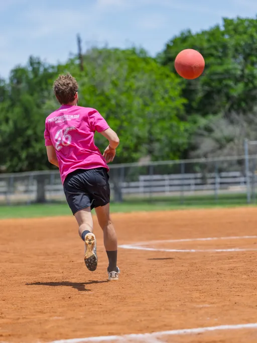 A kickball player wearing a bright pink team shirt and black shorts is running on a dirt kickball field, watching the kickball as it flies through the air. The field is outdoors with a fence and trees in the background.