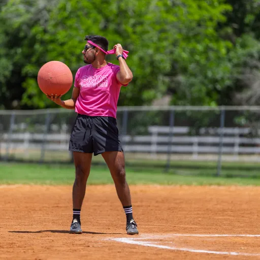 A kickball player in a pink shirt, black shorts, and a matching pink headband prepares to throw a kickball on a sunny outdoor field. The athlete stands near the pitcher's mound, looking toward their target with teammates and trees in the background.