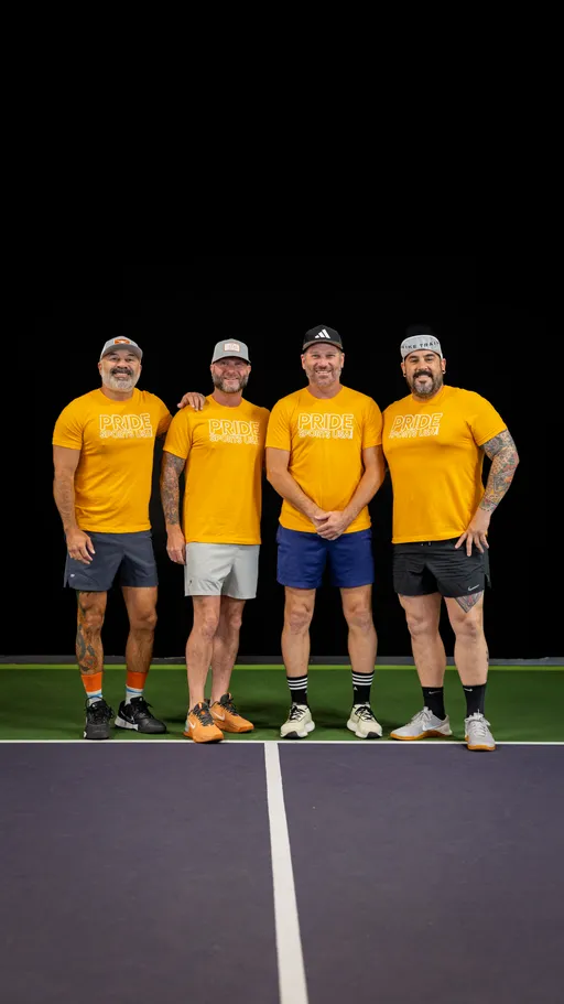 Four pickleball players stand side by side on an indoor court, all wearing matching yellow "Pride Sports USA" t-shirts, athletic shorts, and sneakers, with some players also wearing caps and visible tattoos.