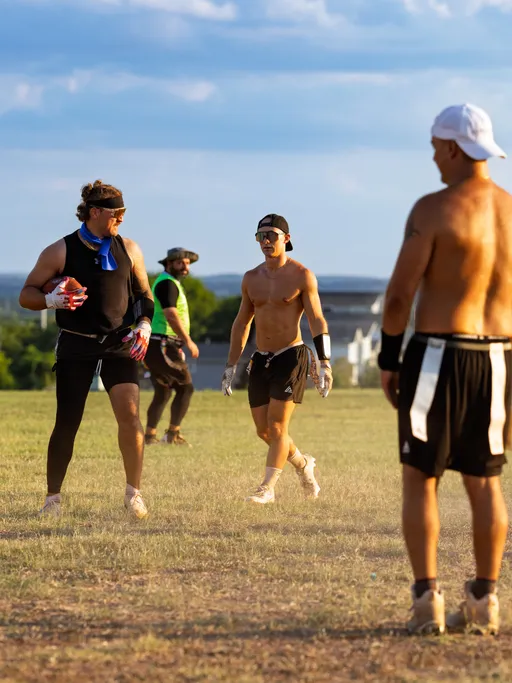 ATX Heat (Black) athletes run flag-football drills on a sunlit grassy field for Gay Bowl 2025 practice: one player in black shorts and receiver gloves carries a red football while teammates wearing flag belts, caps or headbands, and cleats jog and reset between reps against a distant skyline.