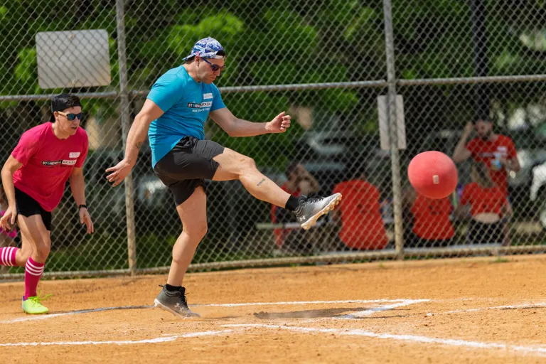 A player in a blue shirt and bandana is kicking a red kickball on a dirt field while a competitor in a pink shirt and striped socks runs behind; a chain-link fence and spectators in the background indicate an outdoor kickball game.
