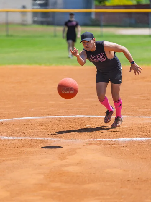 A kickball player wearing a black "Pride Sports" shirt, pink knee-high socks, and a black cap is in mid-motion, pitching a red kickball on a dirt field, with another player visible in the background.