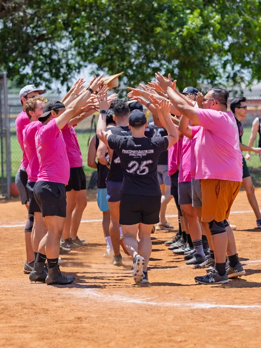 Players in bright pink shirts form a tunnel with their arms raised as another player in a black shirt with the number 25 runs underneath on a sunny kickball field.