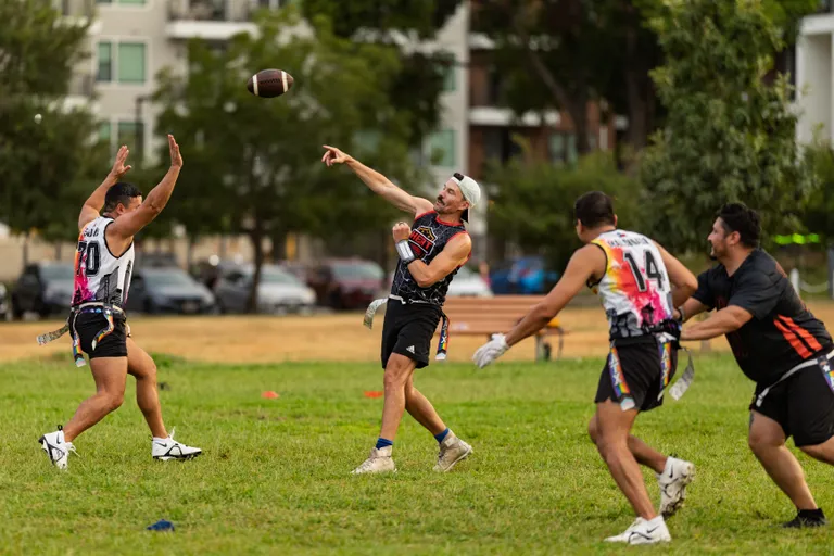ATX Heat (Orange) players scrimmage in a city park: a quarterback in a black jersey with multicolor accents and a backward cap throws a pass as a receiver reaches up and two defenders close in; all wear numbered jerseys, flag belts, and cleats, with cones marking the field.