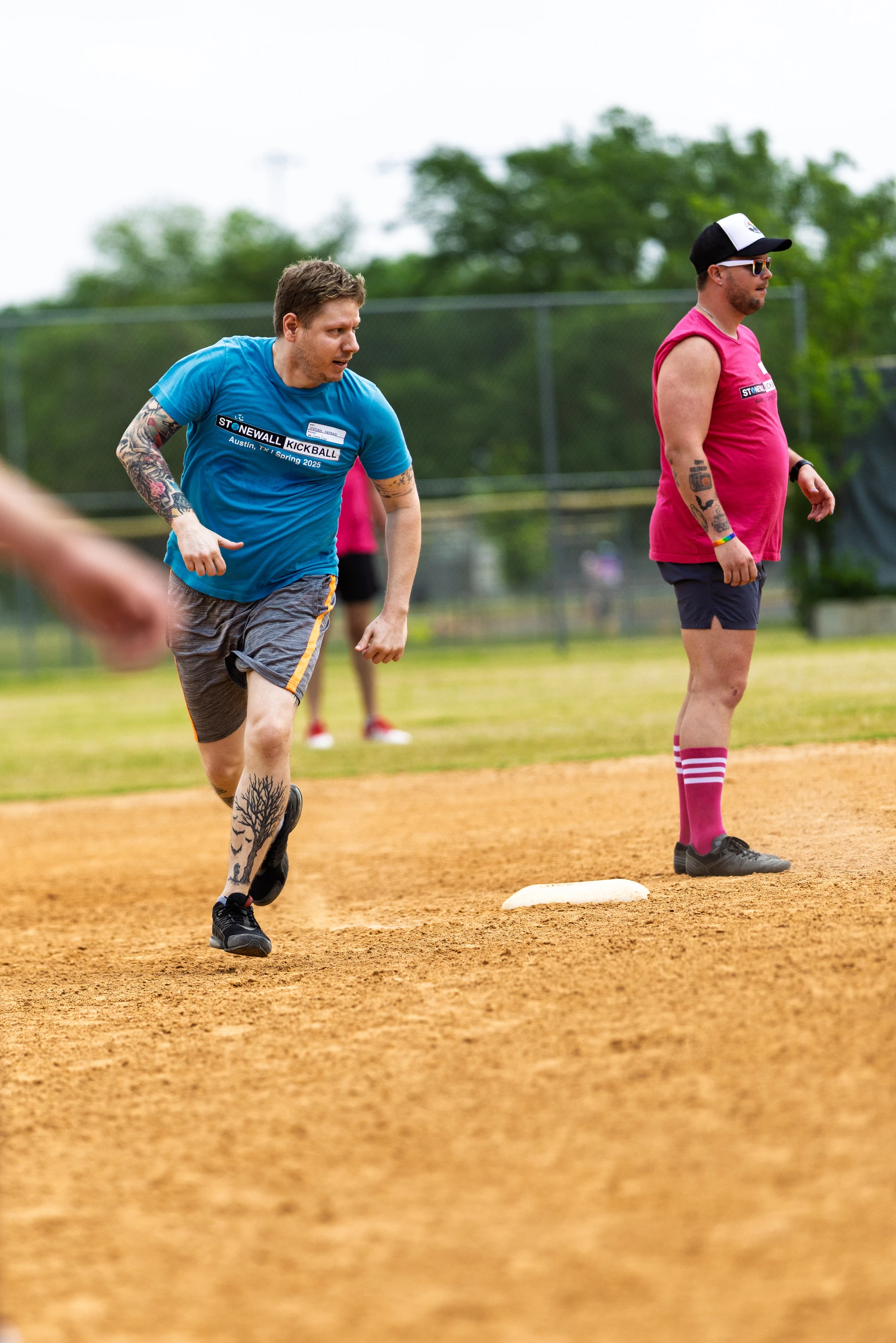 A player wearing a blue "Stonewall Kickball" shirt and shorts is running toward a base on a kickball field, while another player in a pink team shirt, matching socks, and a cap stands near the base watching the play.