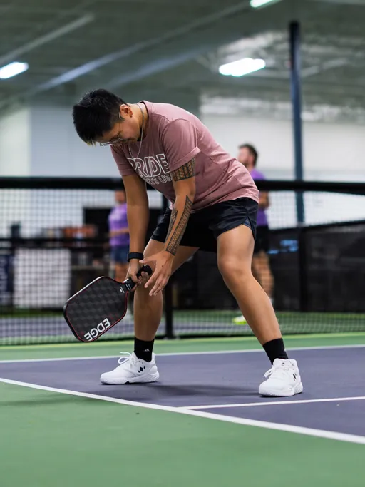 A pickleball player wearing a "PRIDE" t-shirt, shorts, and sports shoes is preparing to serve on an indoor court, holding a paddle and focusing on the ball. The environment includes a visible net and other players in the background.