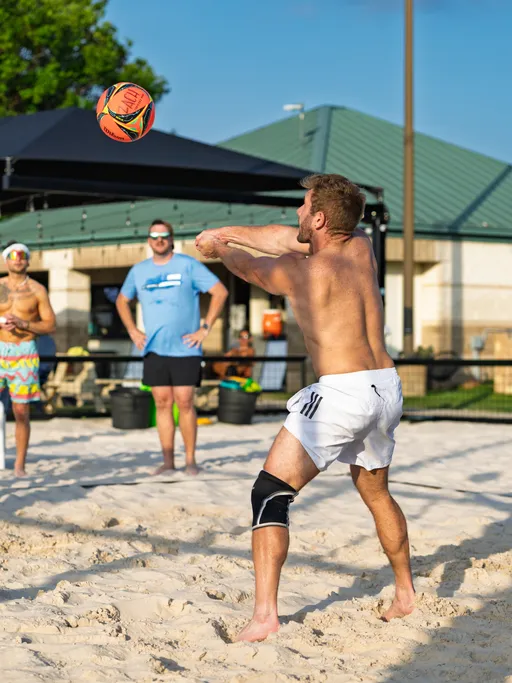 A volleyball player wearing white athletic shorts and a knee brace is preparing to bump a volleyball in a sandy outdoor court, while other players and spectators watch in the background.