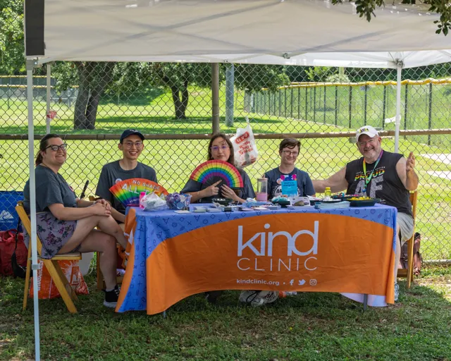 Five people sit at a table under a canopy on a grassy kickball field, with a bright "Kind Clinic" tablecloth and various items on the table, including a colorful fan and drinks; some are smiling and posing for the camera.