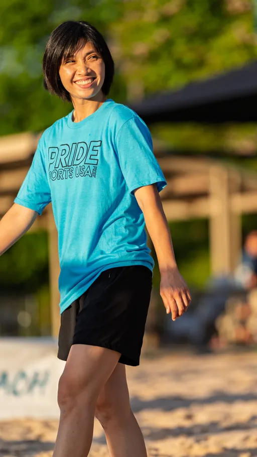 A volleyball player wearing a bright blue "Pride Sports USA" t-shirt and black shorts smiles while walking on a sandy outdoor volleyball court.