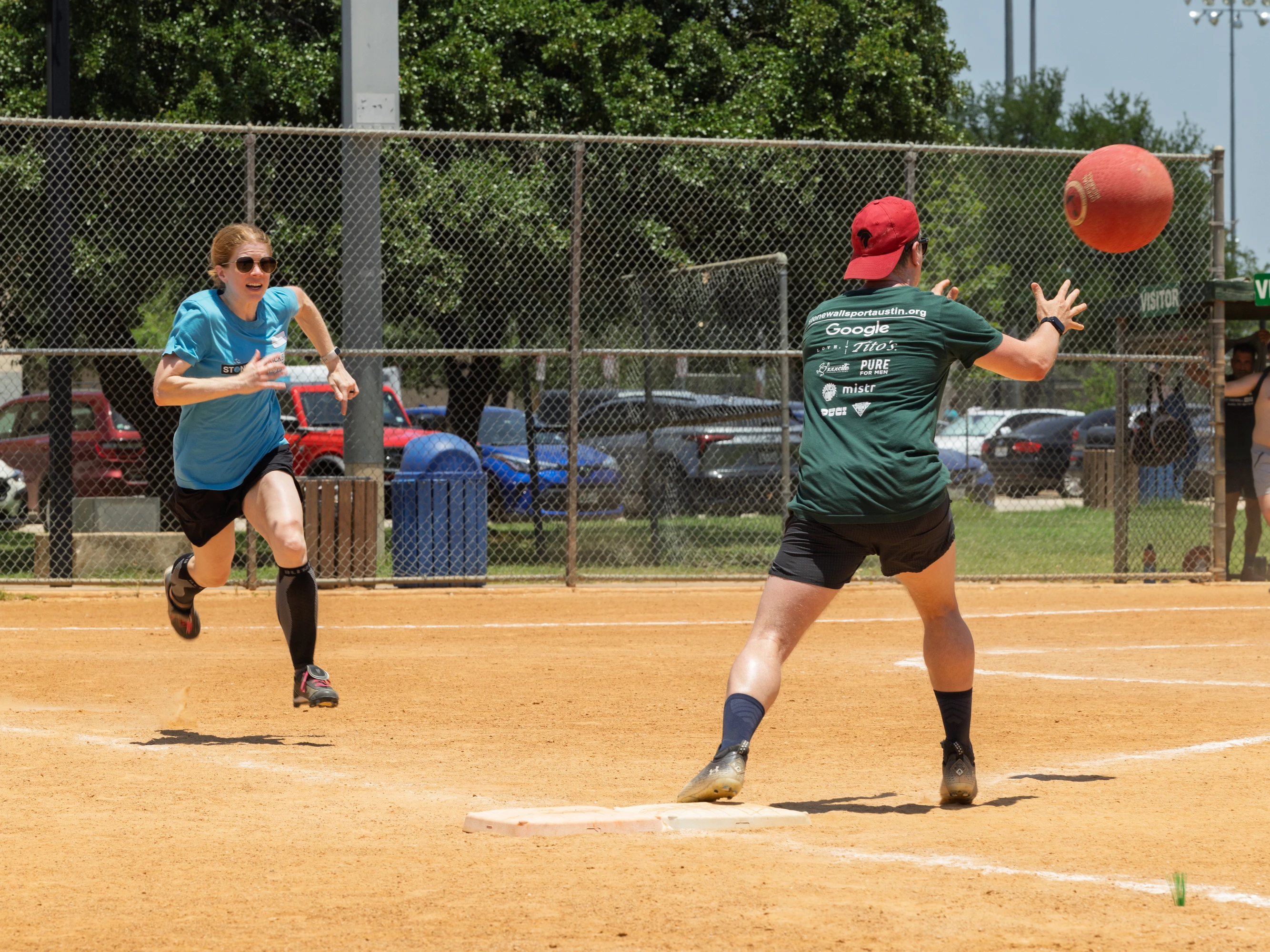 A player wearing a blue shirt and black shorts is running toward a base while another player in a green shirt and red cap prepares to catch a red kickball near the base on a dirt kickball field, with a chain-link fence and parked cars in the background.