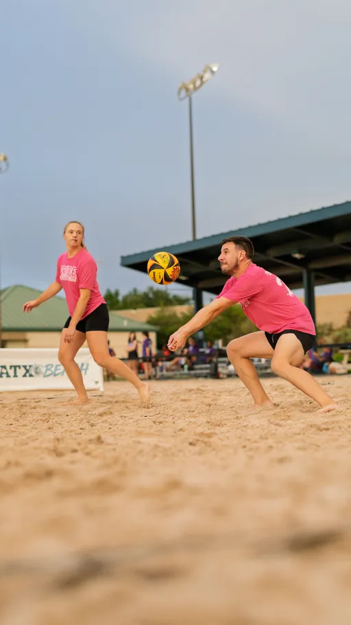 Two volleyball players in matching pink shirts and black shorts are on a sandy court, with one player crouched and arms outstretched to bump the ball while the other watches attentively. Outdoor lights and a covered seating area are visible in the background.