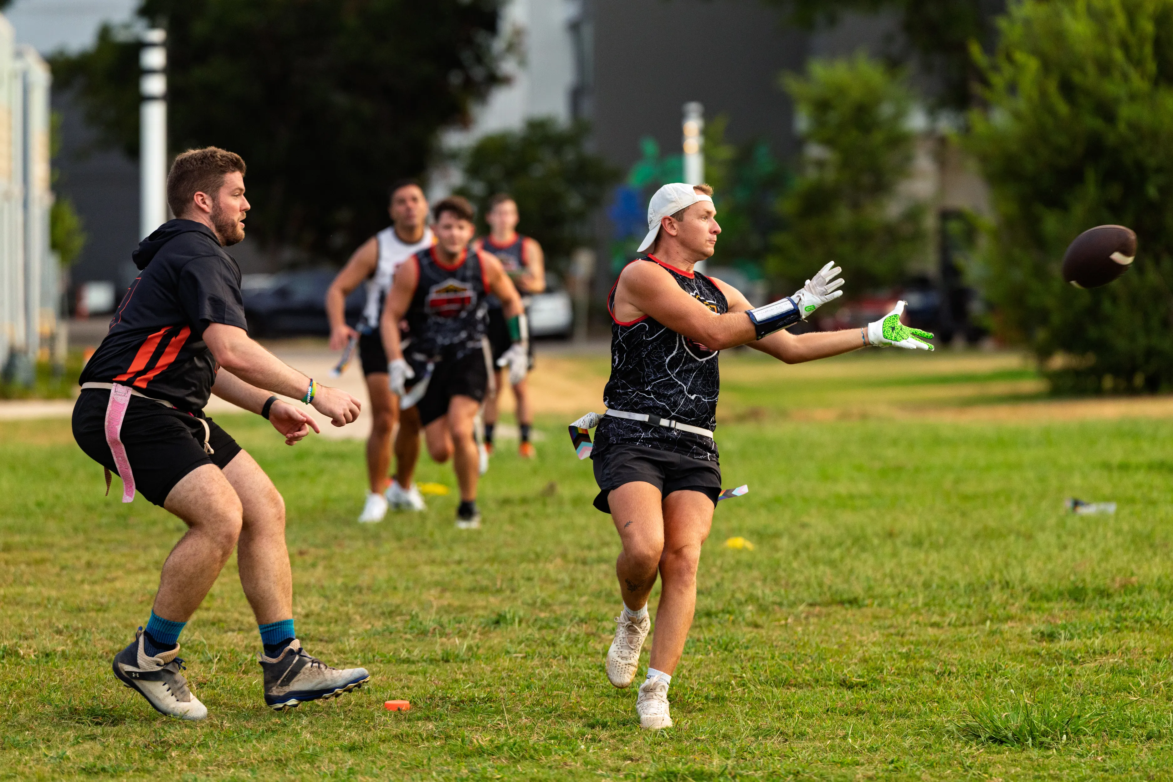 ATX Heat (Orange) athletes scrimmage on a grassy park field as a receiver in a black jersey with orange trim, white cap, gloves, and a flag belt reaches for a pass while a defender closes in. Teammates sprint behind in matching jerseys and cleats, with trees and cones marking the practice area.