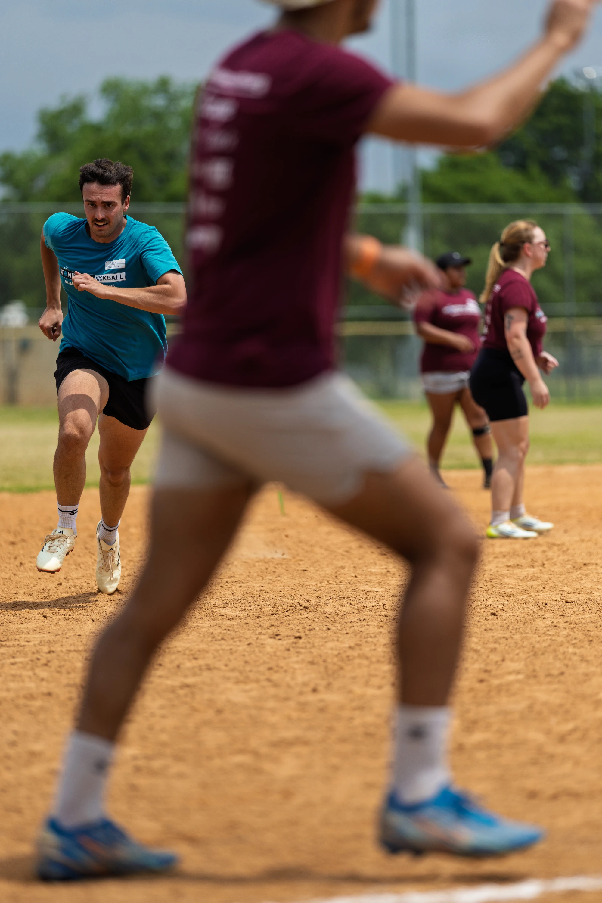 A player in a blue shirt sprints toward a base on a kickball field while another player in a maroon shirt and white shorts stands in the foreground; additional players are visible in the background, all appearing focused on the ongoing game.