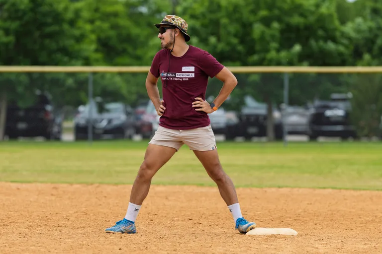 A kickball player stands on a base with hands on hips, wearing a maroon "Stonewall Kickball Spring 2025" shirt, khaki shorts, blue athletic shoes, a patterned bucket hat, and sunglasses on a sandy kickball field with trees and cars in the background.