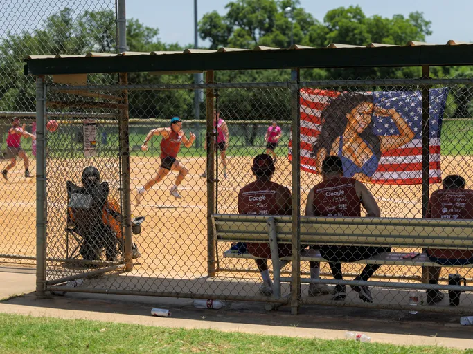 Several kickball players sit in a shaded dugout while another player, wearing a blue hat and orange shirt, runs on the sandy kickball field; a flag with an illustration of a flexing person in front of an American flag is displayed on the fence.