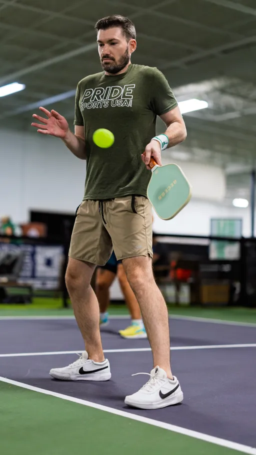 A pickleball player wearing a "Pride Sports USA" T-shirt, shorts, and athletic shoes prepares to hit a ball with a paddle on an indoor pickleball court. The player is focused on the incoming ball during a game.