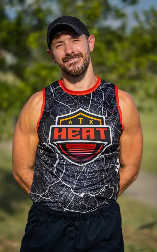 An ATX Heat (Black) athlete stands on a sunny park field during Gay Bowl 2025 practice with hands behind their back, facing forward. The player wears a black cap, dark shorts, and a black sleeveless jersey with red trim and a large “ATX HEAT” crest over a white map-like pattern, with trees and grass blurred in the background.