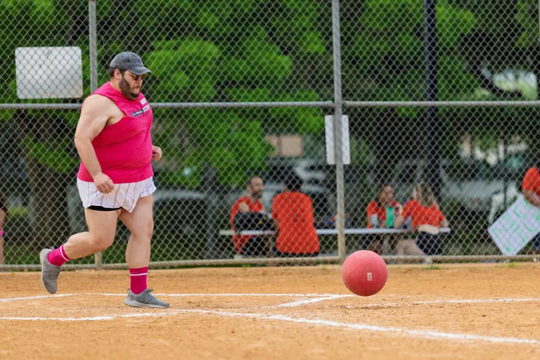 A kickball player wearing a bright pink sleeveless shirt, white shorts, matching pink socks, and a black cap is running toward a red kickball on a dirt field, with a chain-link fence and spectators seated in the background.