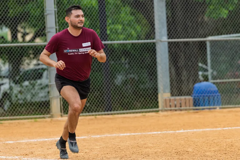 A player wearing a maroon "Stonewall Kickball" t-shirt and black shorts is running on a kickball field, with a chain-link fence and trees in the background.