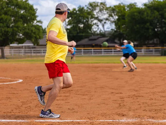 On a sunny kickball field, a player in a yellow shirt and red shorts stands near the baseline watching a green kickball fly toward two fielders in blue shirts who are moving in to catch it. The scene is set on a red-dirt infield with white chalk lines, a fence, and trees in the background.