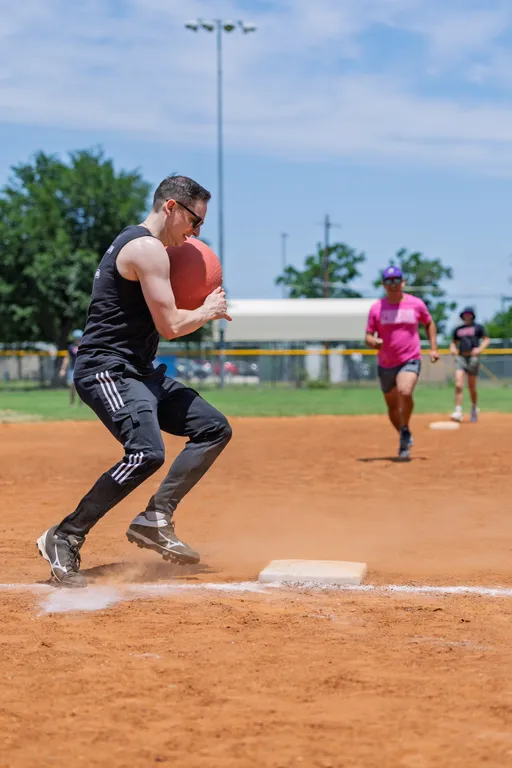 A player in a black tank top, athletic pants, and sunglasses is stepping on a kickball base while clutching a red kickball, with another player in a bright pink shirt running toward the base on a sunny outdoor kickball field.