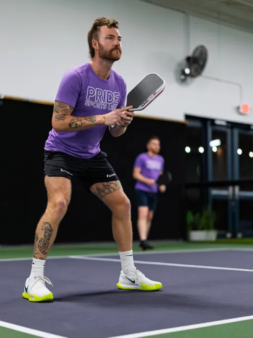 A pickleball player wearing a purple "Pride Sports USA" shirt and black shorts is positioned in an athletic stance, holding a paddle and focused on the game inside an indoor court. Another player stands in the background, also ready for action.
