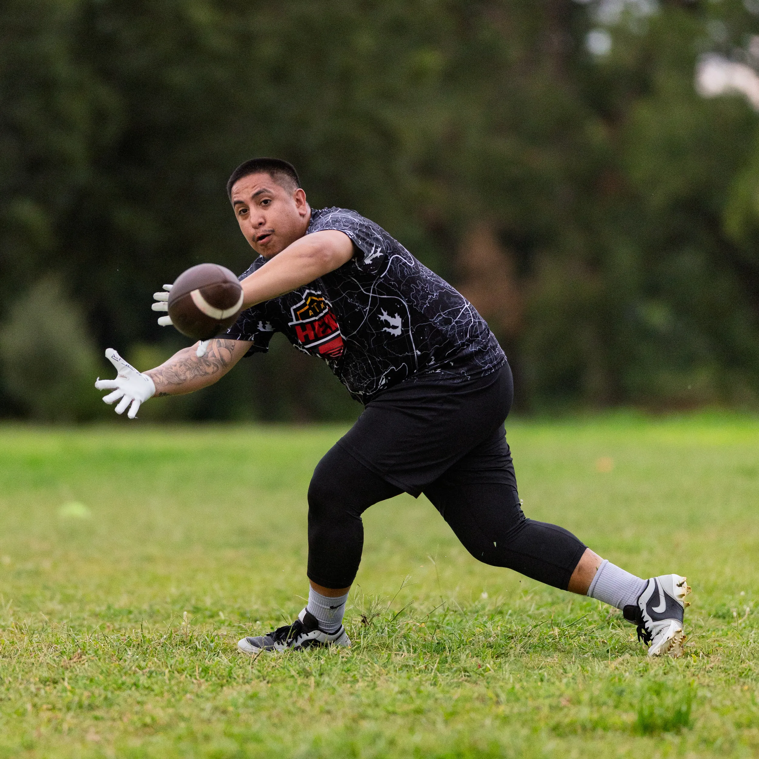 On a grassy field, an ATX Heat flag football player lunges forward with outstretched, gloved hands to snag a low pass during practice, wearing a black patterned team jersey with logo, leggings, socks, and cleats; trees blur in the background.