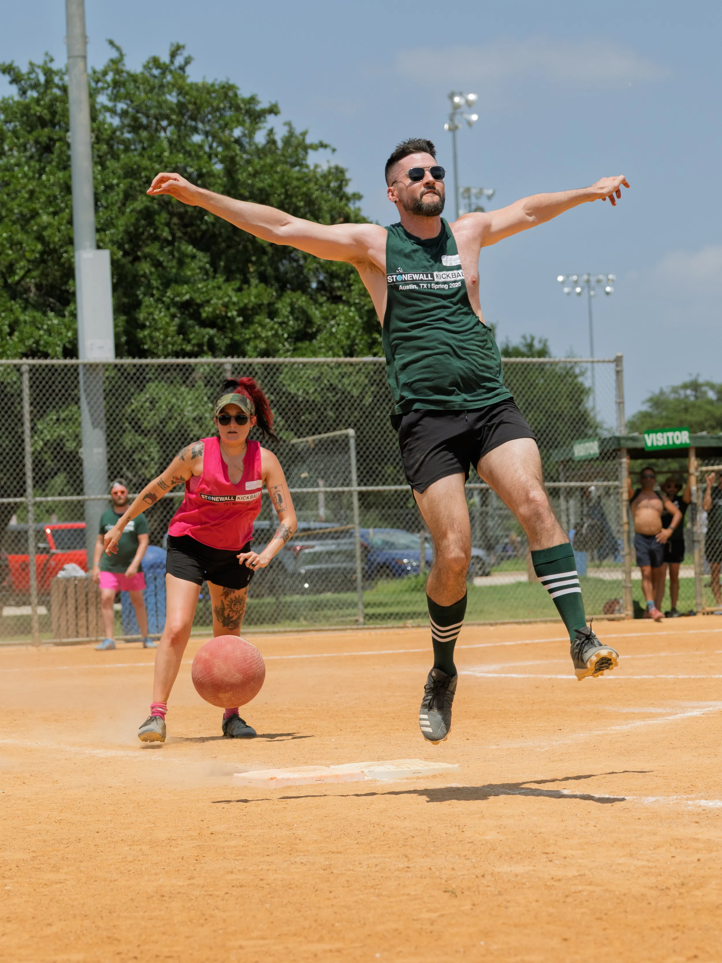On a dusty kickball field, a kicker in a green Stonewall tank, black shorts, and striped knee socks leaps forward mid-swing as a pitcher in a bright pink tank rolls a red kickball toward home plate. Chain-link fencing, trees, and scattered spectators frame the sunny game.