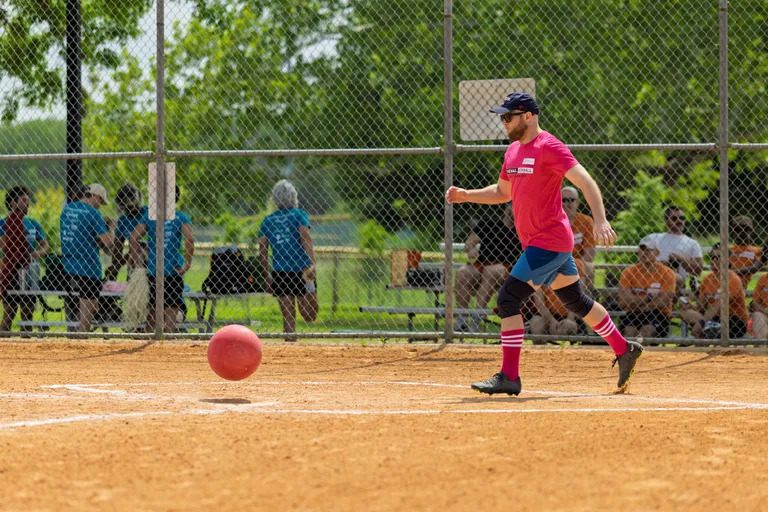 A kickball player wearing a pink shirt, dark shorts, a cap, and knee-high socks is running towards a red kickball on a sandy field, with spectators and other players in the background behind a chain-link fence.