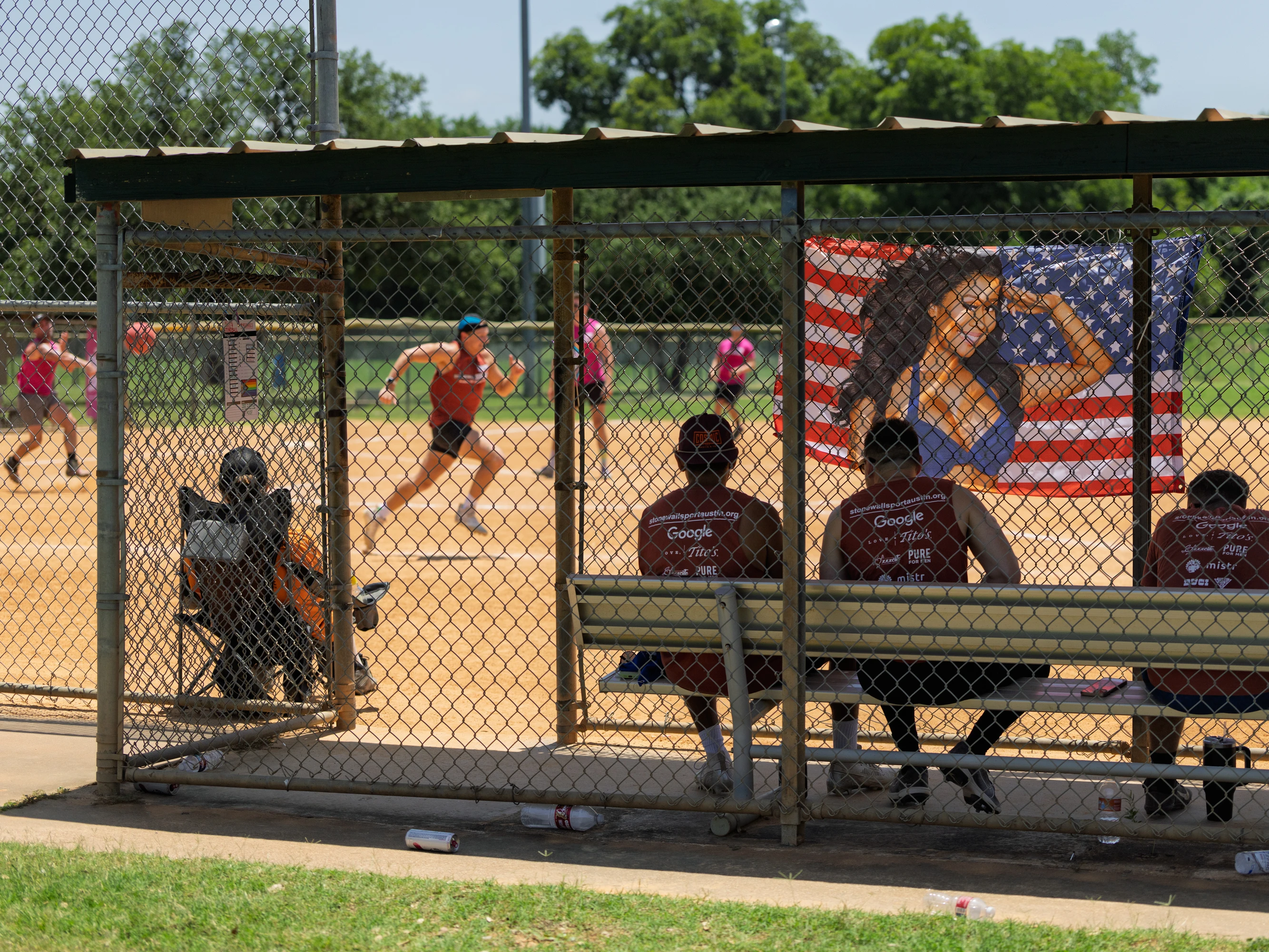 Several kickball players sit in a shaded dugout while another player, wearing a blue hat and orange shirt, runs on the sandy kickball field; a flag with an illustration of a flexing person in front of an American flag is displayed on the fence.