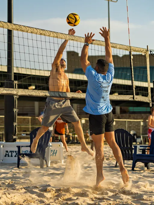 Two volleyball players are jumping at the net; one is attempting to spike or block the ball, while the other is reaching up defensively. Both are on a sandy volleyball court, with one player wearing a light blue shirt and black shorts and the other in shorts without a shirt.