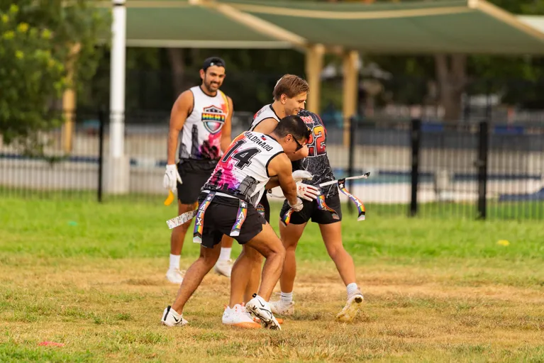 ATX Heat (Orange) players run a flag football drill on a grassy practice field, with one defender reaching to pull a runner’s flag while teammates close in; they wear numbered jerseys with rainbow ATX logos, flag belts, cleats, and some gloves, with shade canopies and a fence in the background.
