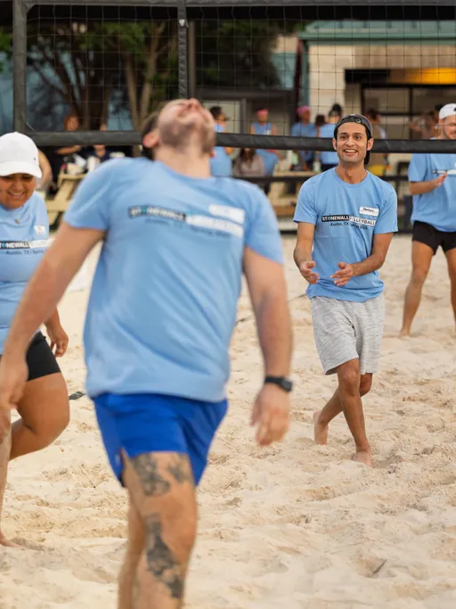 Several volleyball players wearing light blue Stonewall Sand Volleyball shirts and athletic shorts are actively moving on a sand court, with one player laughing and another looking upward, all engaged in gameplay near the net.
